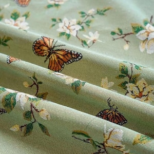 May include: Close-up of a sage green fabric with a repeating pattern of butterflies and floral elements. The butterflies are orange and brown, while the flowers are white and yellow. The fabric has a textured appearance.