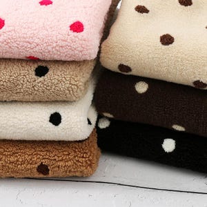 Könnte beinhalten: Ein Stapel weicher, strukturierter Stoffe in verschiedenen Farben, darunter Rosa, Beige, Weiß, Braun und Schwarz. Jedes Stück hat ein Tupfenmuster in Kontrastfarben. Der Stoff wirkt weich und gemütlich, geeignet für Decken oder Kleidung.