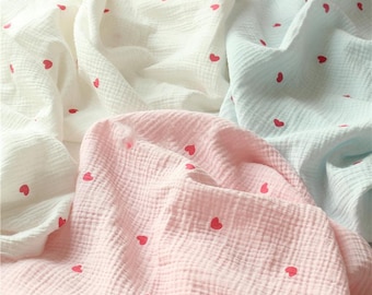 May include: Close-up of soft, textured fabric in white, pink, and light blue. The fabric is adorned with small, evenly spaced red hearts. The material appears to be lightweight and suitable for baby blankets or other delicate items.
