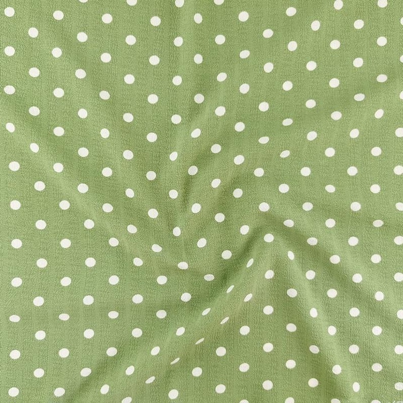 May include: A sage green fabric featuring a repeating pattern of small, evenly spaced white polka dots. The material has a subtle, crinkled texture with soft folds that create gentle shadows across the surface of the textile.