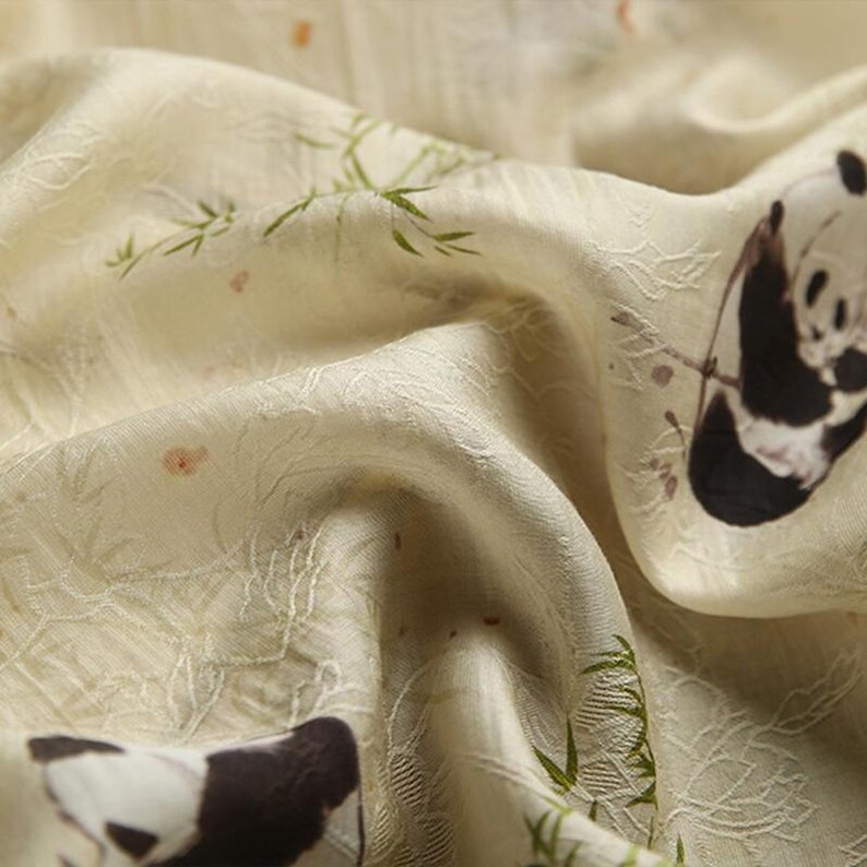 Pu&ograve; includere: Un tessuto color crema caratterizzato da un motivo ripetuto di panda bianchi e neri, steli di bamb&ugrave; verdi e piccoli accenti arancioni. Il materiale presenta una superficie testurizzata e leggermente stropicciata con un motivo intrecciato.