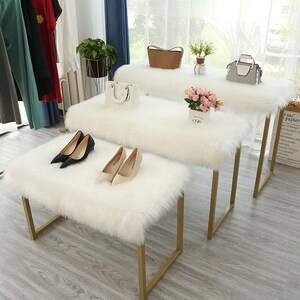 May include: Three rectangular benches with white faux fur tops and gold metal frames arranged in a tiered formation. The benches display various items including black and beige high-heeled shoes, a white handbag, a brown handbag, a gray handbag, and a small potted plant with pink flowers.