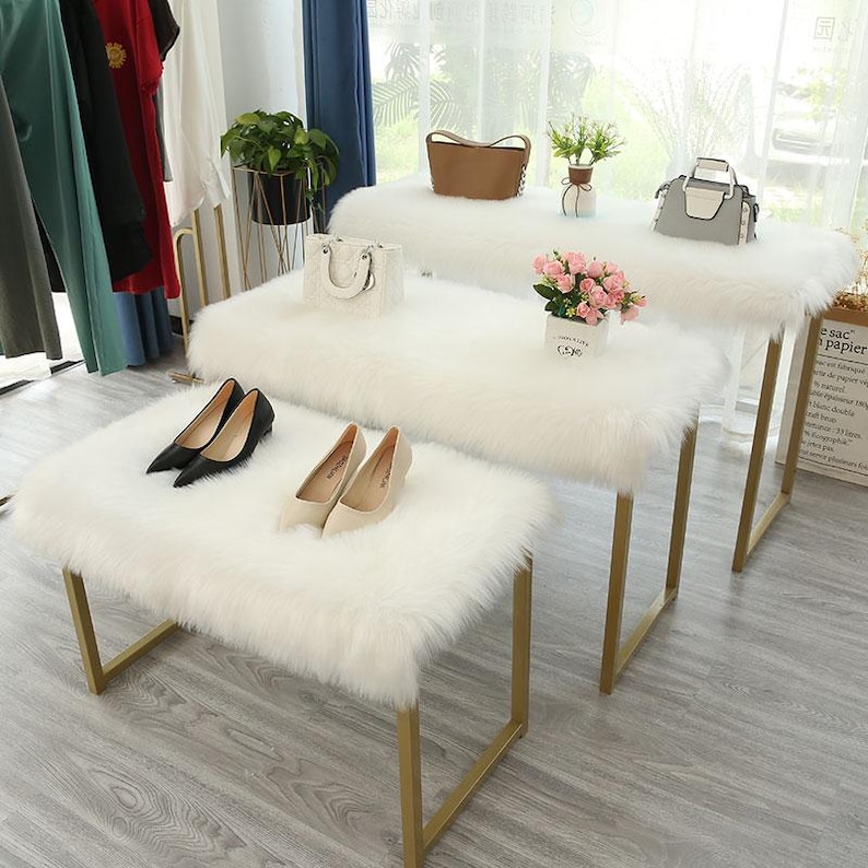 May include: Three rectangular benches with white faux fur tops and gold metal frames arranged in a tiered formation. The benches display various items including black and beige high-heeled shoes, a white handbag, a brown handbag, a gray handbag, and a small potted plant with pink flowers.