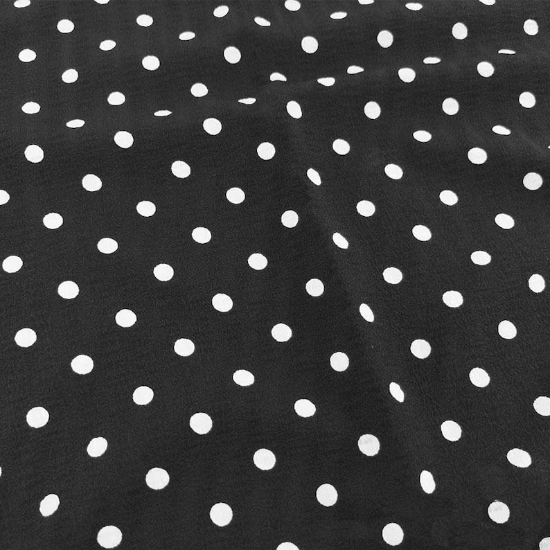 May include: A black fabric featuring a repeating pattern of small, solid white polka dots distributed evenly across the surface. The material displays soft folds and gentle ripples, creating a textured appearance with light and shadow variations.