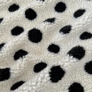 Könnte beinhalten: Ein dicker, cremefarbener Sherpa-Fleece-Stoff mit einem sich wiederholenden Muster aus großen, soliden schwarzen kreisförmigen Punkten. Das Material hat eine weiche, flauschige und strukturierte Oberfläche mit sichtbaren Flor-Schlingen.