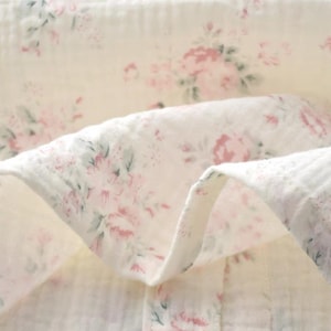 May include: A soft, off-white muslin fabric featuring a delicate floral pattern with muted pink roses and sage green leaves. The textile has a crinkled, multi-layered texture with visible folds that create depth and highlight the floral print design.