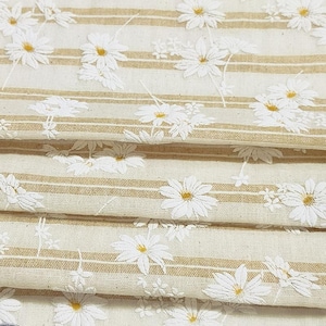May include: A folded piece of fabric featuring a pattern of white daisy flowers with yellow centers set against a background of alternating cream-colored sections and thin, horizontal tan stripes running across the textile surface.