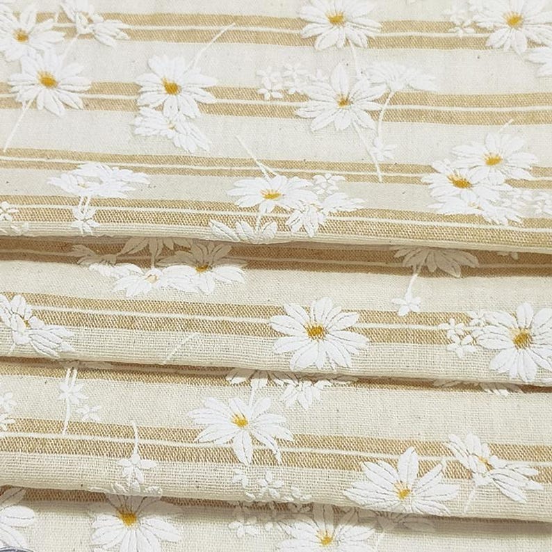 May include: A folded piece of fabric featuring a pattern of white daisy flowers with yellow centers set against a background of alternating cream-colored sections and thin, horizontal tan stripes running across the textile surface.