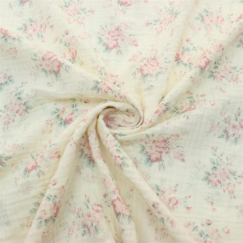May include: A soft, off-white muslin fabric featuring a repeating floral pattern of muted pink roses and sage green leaves. The textile has a crinkled, multi-layered texture with a gathered center point that creates a swirling, dimensional effect.