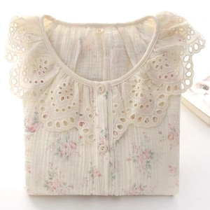 May include: A cream-colored button-down shirt featuring a delicate pink floral pattern and a wide, scalloped eyelet lace collar. The fabric has a textured, crinkled appearance, and the interior neck label clearly displays the size M.