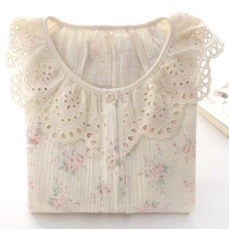 May include: A cream-colored button-down shirt featuring a delicate pink floral pattern and a wide, scalloped eyelet lace collar. The fabric has a textured, crinkled appearance, and the interior neck label clearly displays the size M.