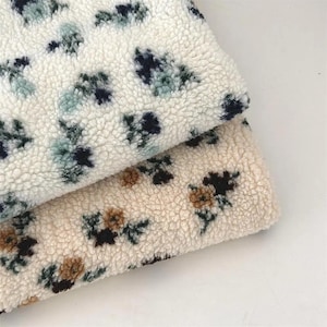 May include: Close-up of two pieces of soft, textured fabric. One is off-white with blue and green floral patterns, and the other is beige with brown and black floral patterns. The fabric appears to be a cozy, plush material.