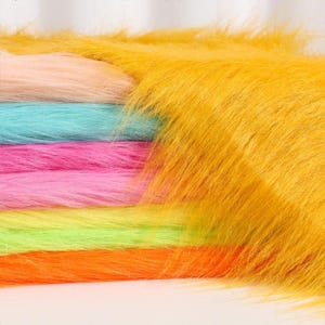 May include: A stack of long-pile faux fur fabric in vibrant colors including mustard yellow, peach, turquoise, hot pink, lime green, and bright orange. Each layer features a soft, shaggy texture with long, flowing synthetic fibers.