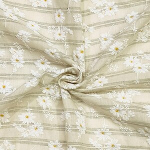 May include: A beige fabric featuring a pattern of white embroidered daisies with yellow centers. The textile is decorated with thin, horizontal sage green stripes running across the surface, creating a textured and layered appearance.