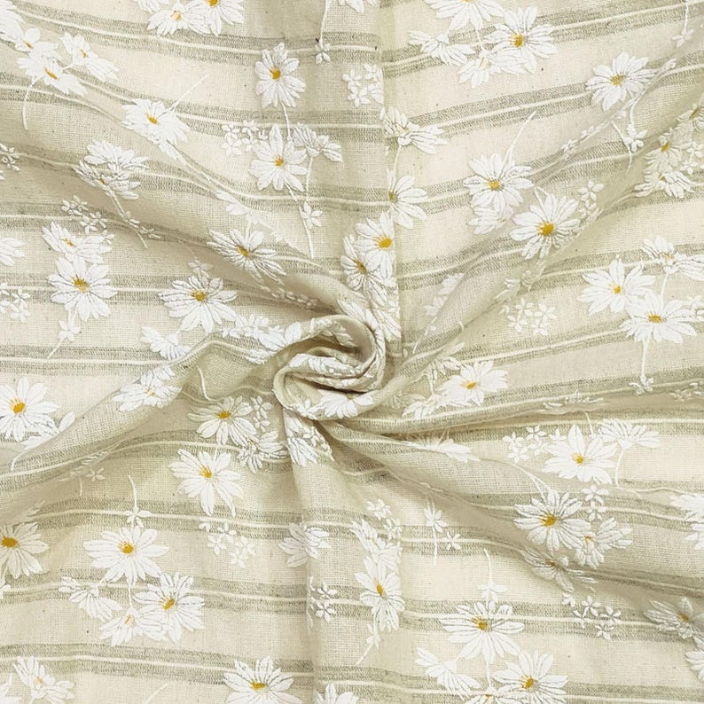 May include: A beige fabric featuring a pattern of white embroidered daisies with yellow centers. The textile is decorated with thin, horizontal sage green stripes running across the surface, creating a textured and layered appearance.