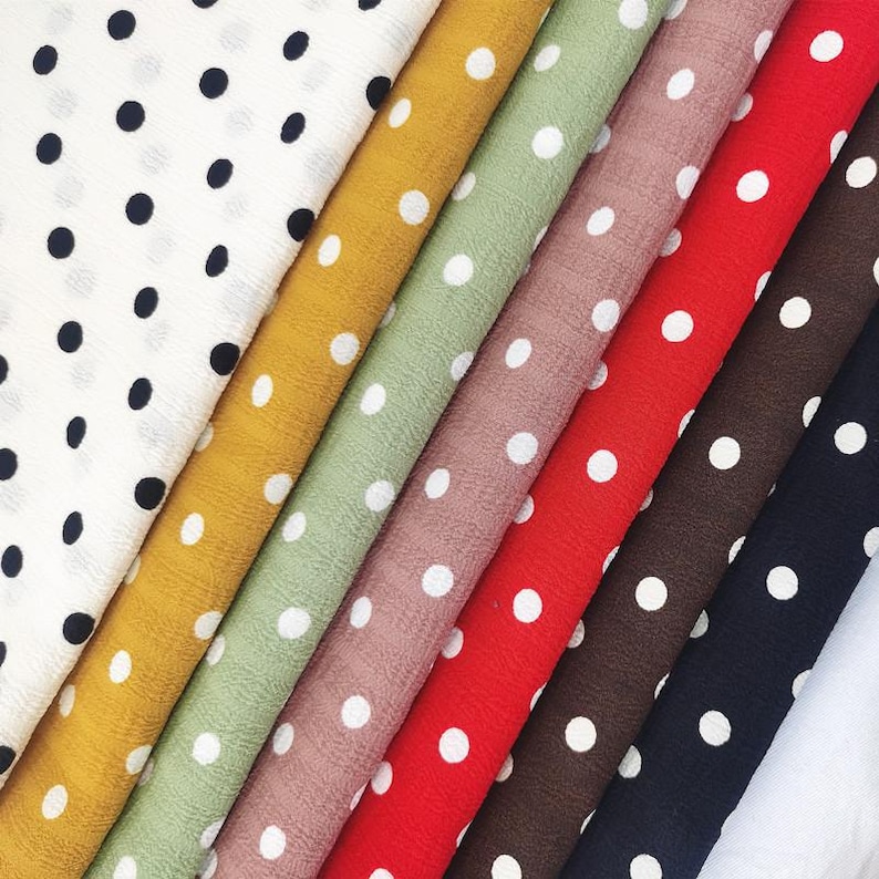 May include: A collection of folded fabric swatches arranged diagonally. The fabrics feature a consistent white polka dot pattern on various solid background colors including white, mustard yellow, sage green, dusty rose, red, brown, and black.