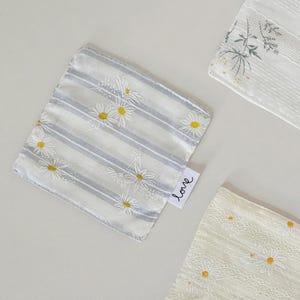 May include: Three square fabric cloths featuring white daisy patterns with yellow centers. One cloth has grey stripes, while the others have textured white fabric. A small white tag on the striped cloth displays the word love in black script.