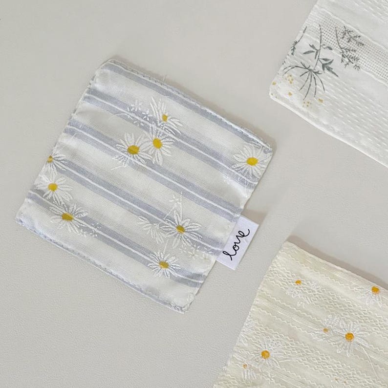 May include: Three square fabric cloths featuring white daisy patterns with yellow centers. One cloth has grey stripes, while the others have textured white fabric. A small white tag on the striped cloth displays the word love in black script.