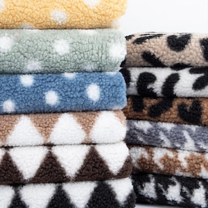 May include: A stack of different patterned fleece fabrics. The fabrics feature polka dots, leopard print, and geometric designs in various colors, including beige, brown, blue, and green.