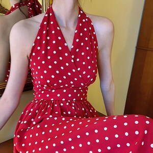 May include: A red halter-neck dress featuring a white polka dot pattern throughout. The garment has a V-neckline, a fitted waistband, and a flowing skirt. The design includes a wrap-style bodice that secures at the neck, creating an open back.