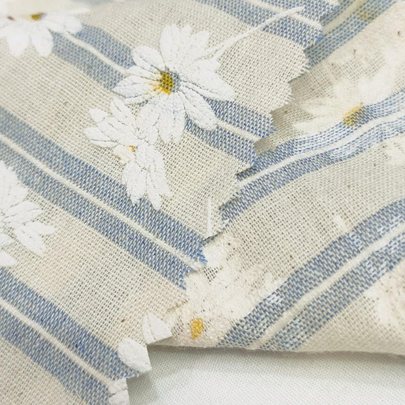 May include: A piece of woven fabric featuring a light beige background with vertical blue stripes and a repeating pattern of white daisy flowers with yellow centers. The material has a visible textured weave and frayed edges along the cut sections.