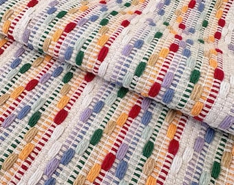 Puede incluir: Un tejido con una base de color crema y rayas verticales de hilo elevado y copetudo en varios colores, incluyendo rojo, verde, azul, amarillo y lavanda. La tela presenta una apariencia texturizada y multicolor con un patrón repetitivo.