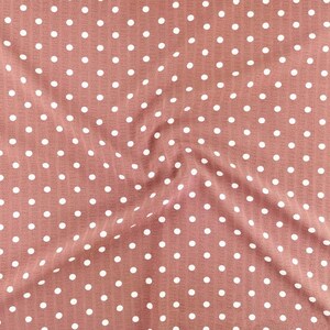 May include: A dusty rose pink fabric featuring a repeating pattern of small white polka dots. The material has a subtle vertical ribbed texture throughout, with soft folds creating natural shadows and highlights across the surface of the textile.