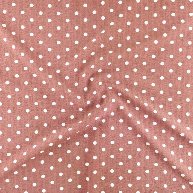 May include: A dusty rose pink fabric featuring a repeating pattern of small white polka dots. The material has a subtle vertical ribbed texture throughout, with soft folds creating natural shadows and highlights across the surface of the textile.