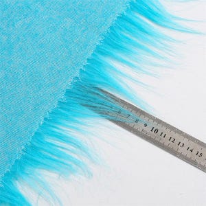 May include: A bright turquoise fabric featuring a long, shaggy faux fur trim along one edge. A metal ruler is placed over the fur, showing the length of the fibers extending from the edge of the fabric to approximately 2.5 inches in length.