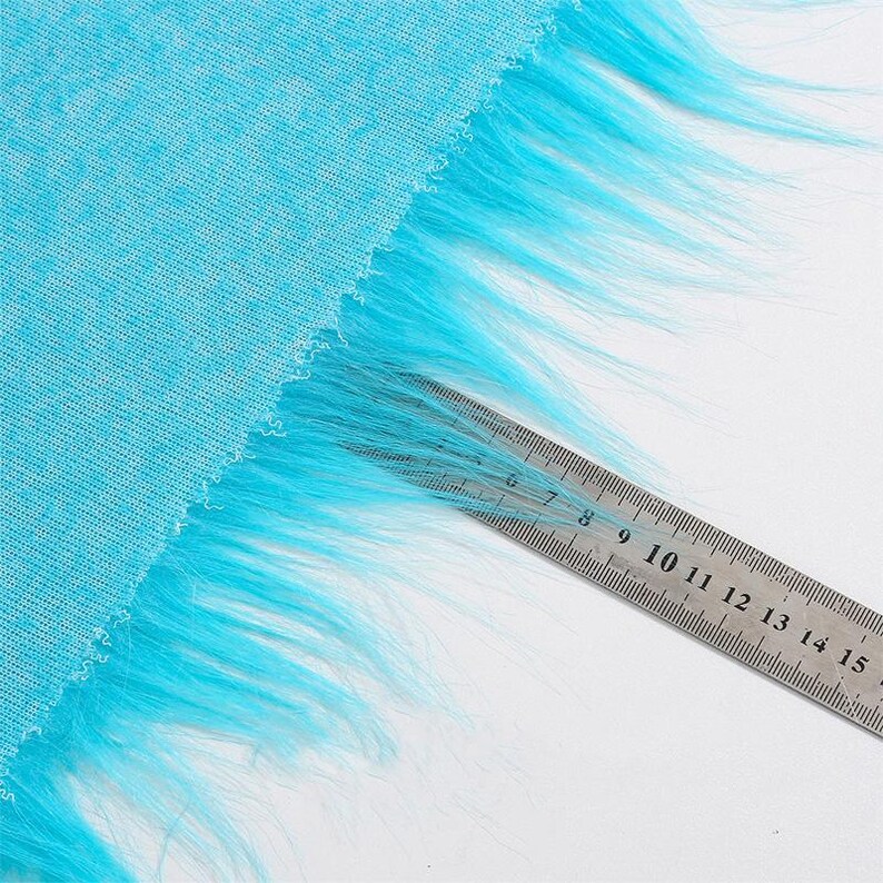 May include: A bright turquoise fabric featuring a long, shaggy faux fur trim along one edge. A metal ruler is placed over the fur, showing the length of the fibers extending from the edge of the fabric to approximately 2.5 inches in length.