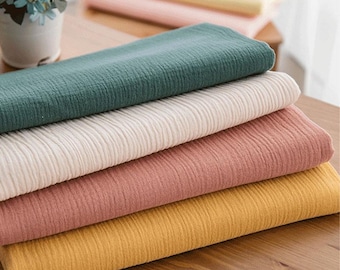 May include: A stack of folded fabric in various colors: teal, cream, dusty rose, and mustard yellow. The fabric has a textured, crinkled appearance. The image is taken from a slightly elevated angle, showing the layers of fabric.