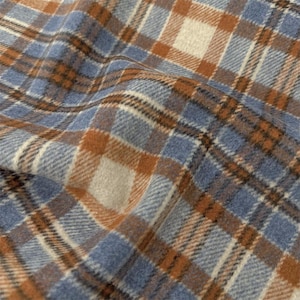 Puede incluir: Una tela tejida con un patrón de cuadros en tonos azul polvoriento, marrón cálido y blanco roto. El material tiene una textura suave y ligeramente difusa con líneas diagonales visibles y un diseño de cuadrícula repetitivo en toda la superficie.