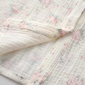 May include: A cream-colored muslin fabric shirt featuring a delicate floral pattern in shades of pink and muted green. The garment has a button-down front with a visible button, a folded collar, and a soft, crinkled texture throughout the material.