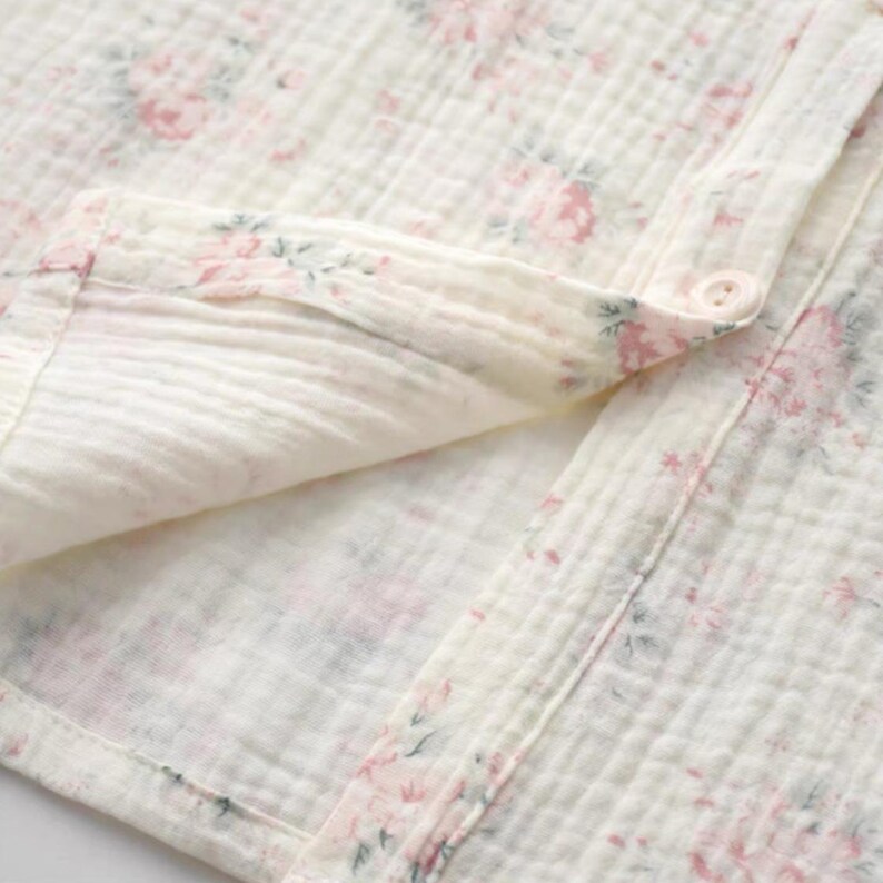 May include: A cream-colored muslin fabric shirt featuring a delicate floral pattern in shades of pink and muted green. The garment has a button-down front with a visible button, a folded collar, and a soft, crinkled texture throughout the material.