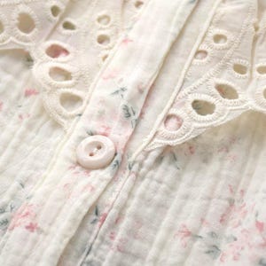 May include: A cream-colored button-down shirt featuring a delicate pink and green floral print on a textured fabric. The garment includes a wide collar with intricate white eyelet embroidery and a single round, light-colored plastic button.