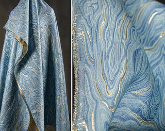 May include: A length of fabric featuring a textured, swirling marble pattern in shades of light and dark blue, accented with metallic gold thread detailing. The material has a visible weave and a frayed edge, showcasing the intricate, flowing design.