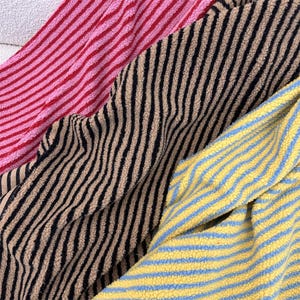 Op de afbeelding: Drie stukken gestructureerde fleecestof met strepenpatronen. Eén stuk heeft roze en rode strepen, een ander heeft beige en zwarte strepen en het derde stuk toont gele en lichtblauwe strepen. Het materiaal heeft een zacht, pluizig oppervlak.