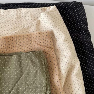 May include: A selection of fabric samples in shades of black, cream, tan, and olive green. Each fabric features a small, repeating white dot design. The fabrics are arranged in layers, highlighting their textures and colours.