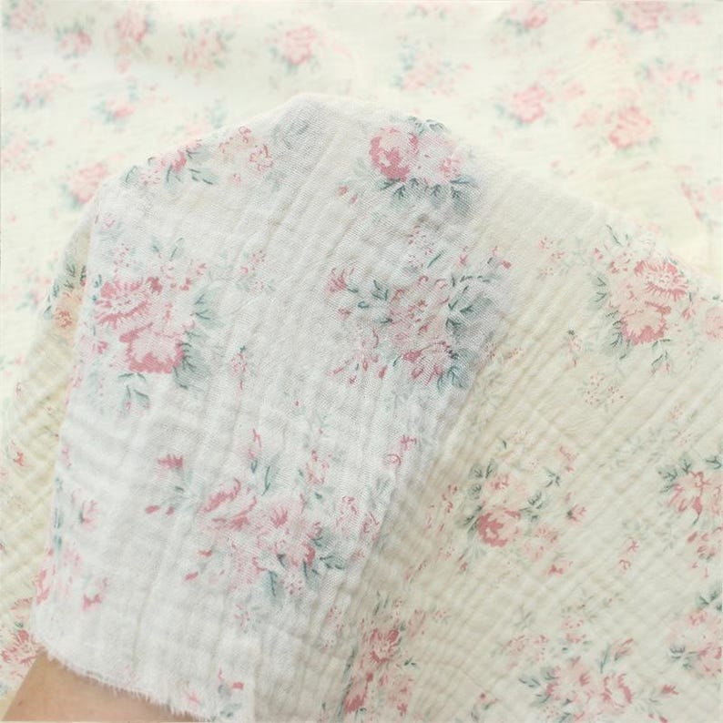 May include: A piece of off-white double gauze fabric featuring a repeating floral pattern in soft pink and muted sage green. The textile has a distinct crinkled, puckered texture characteristic of muslin, with visible raw edges and a lightweight weave.