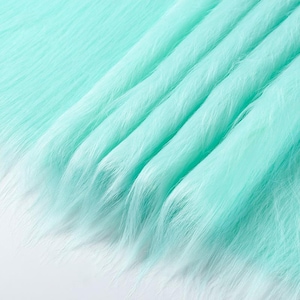May include: A length of mint green faux fur fabric featuring long, soft synthetic fibers. The material is draped in gentle, flowing folds, creating a textured surface with a consistent light green color throughout the entire piece of textile.