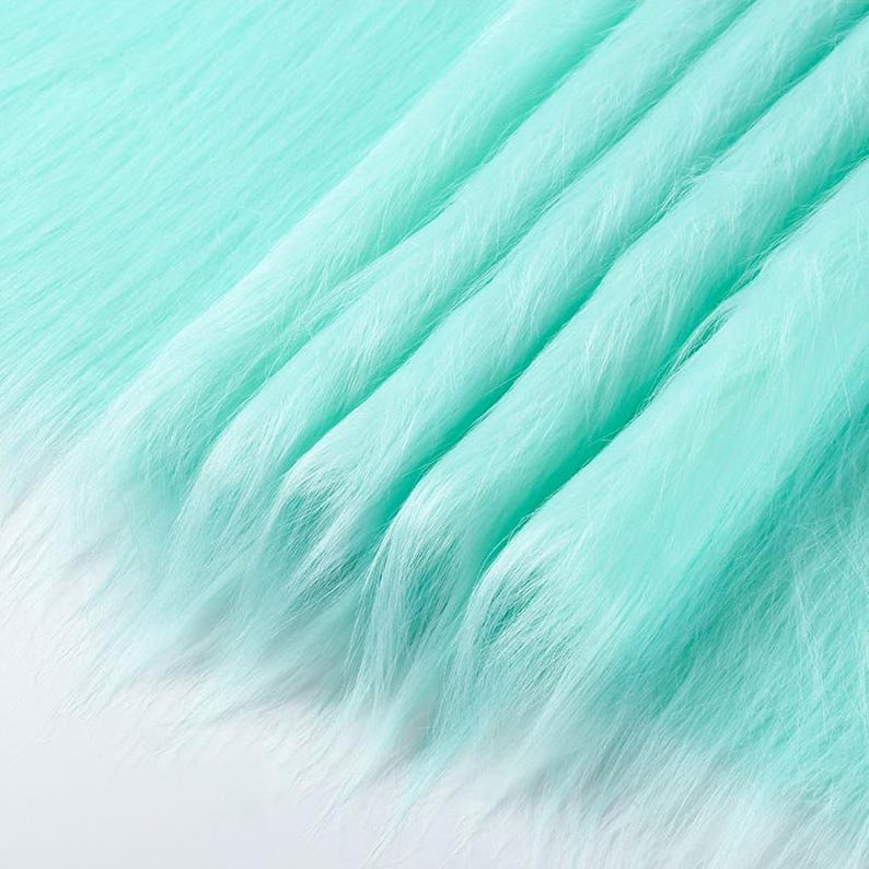 May include: A length of mint green faux fur fabric featuring long, soft synthetic fibers. The material is draped in gentle, flowing folds, creating a textured surface with a consistent light green color throughout the entire piece of textile.
