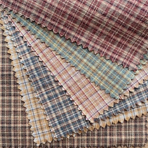 Può includere: Una collezione di campioni di tessuto con vari motivi a quadri in tonalità rosso scuro, blu tenue, giallo dorato e marrone. Ogni campione presenta bordi dentellati ed è sovrapposto per mostrare le diverse texture intrecciate.