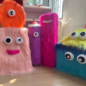 May include: A collection of five plush monster-shaped items covered in long faux fur. The group includes a round orange piece, a tall pink rectangular piece with a winking face, a purple cylinder, a tall magenta piece with multiple eyes, and a blue and yellow block.
