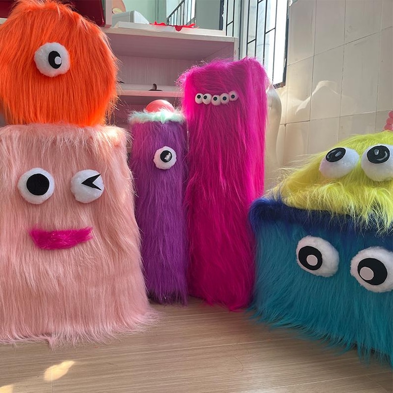 May include: A collection of five plush monster-shaped items covered in long faux fur. The group includes a round orange piece, a tall pink rectangular piece with a winking face, a purple cylinder, a tall magenta piece with multiple eyes, and a blue and yellow block.