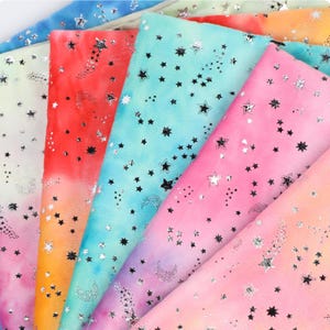 May include: Multiple folded pieces of soft fabric featuring a vibrant watercolour-style gradient in shades of blue, pink, orange, and yellow. Each piece is decorated with a repeating pattern of small, metallic silver stars and crescent moons.