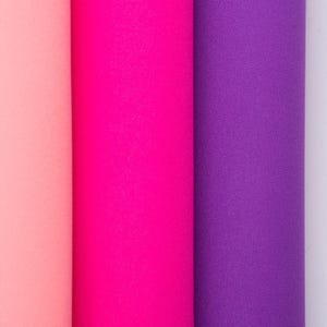 May include: Three vertical rolls of fabric displayed side by side. The first roll is a soft pastel peach colour, the second roll is a vibrant neon pink, and the third roll is a deep, saturated purple. Each fabric roll has a consistent texture.