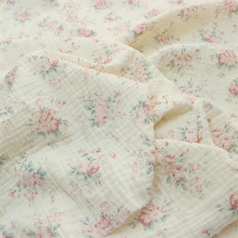 May include: A soft, off-white muslin fabric featuring a repeating floral pattern of muted pink roses and sage green leaves. The textile has a distinct crinkled, multi-layered texture with visible folds and gathers throughout the draped material.