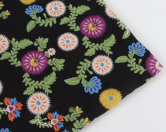 May include: A black fabric featuring a dense, colorful floral embroidery pattern. The design includes circular blossoms in shades of purple, yellow, and white, alongside blue and orange flowers with green leafy stems and vines throughout.