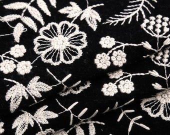 May include: A black fabric featuring an intricate white embroidered pattern of various flowers, leaves, and stems. The embroidery consists of detailed floral motifs and foliage arranged across the dark textile surface in a repeating design.