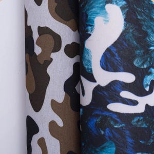 May include: Two rolls of fabric displayed side by side. The left roll features a traditional camouflage pattern in shades of brown, black, and white. The right roll displays a vibrant blue and black abstract camouflage pattern with white accents.